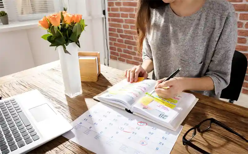 Person writing in diary planner at a desk with a laptop