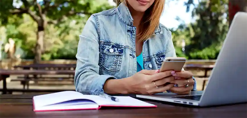 Person sitting at park table looking at their smart phone and a laptop and notebook by their side
