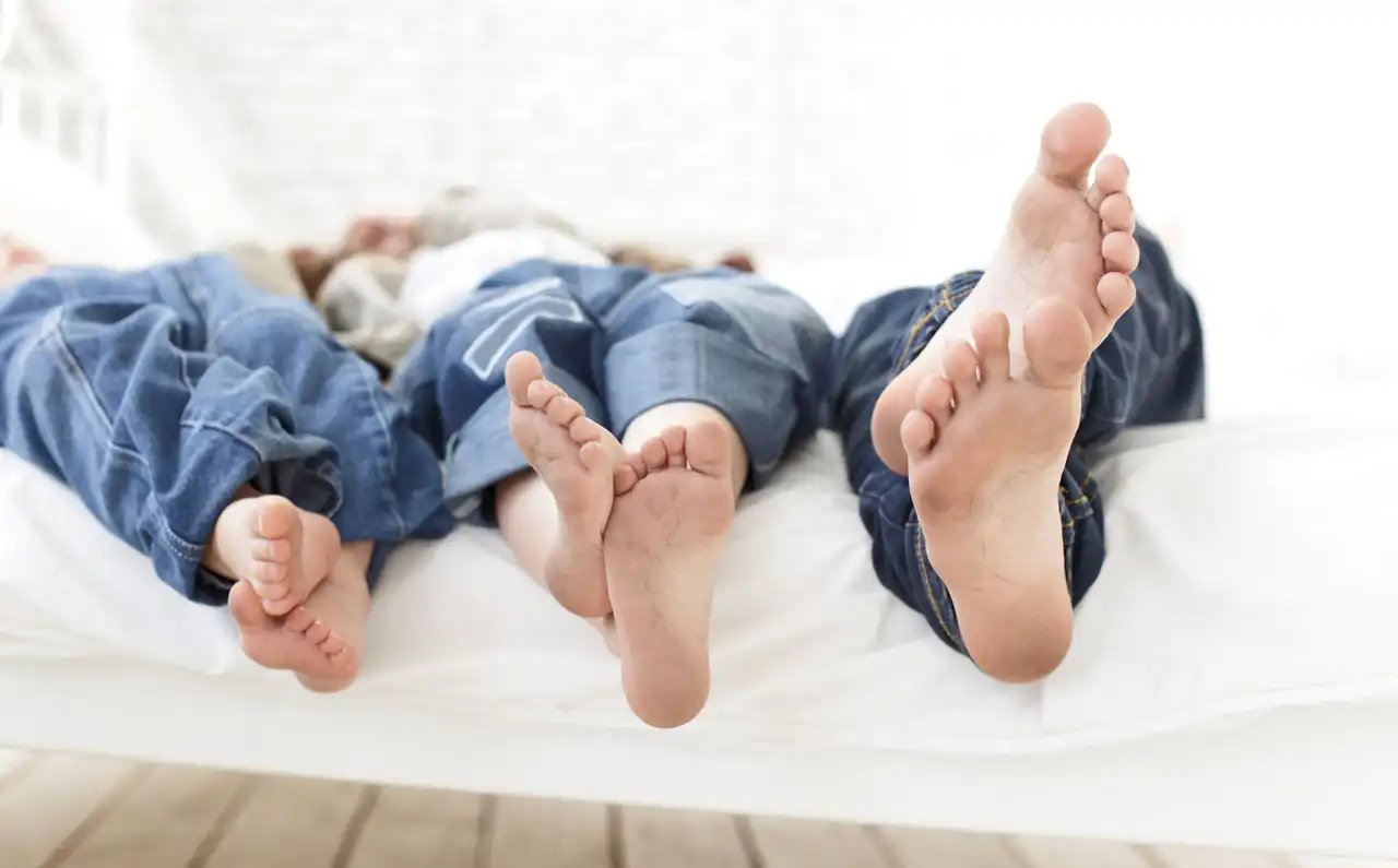 Feet of three people lying on a bed in blue jeans with no shoes or socks