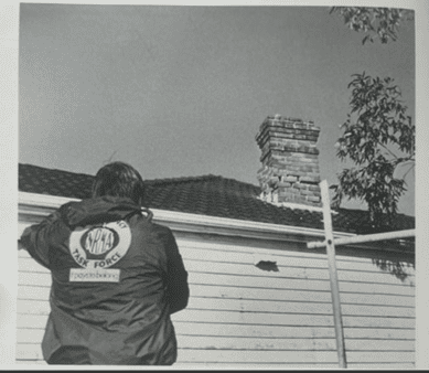 Black and white photo from 1989 of a person wearing an NRMA jacket, facing a house.
