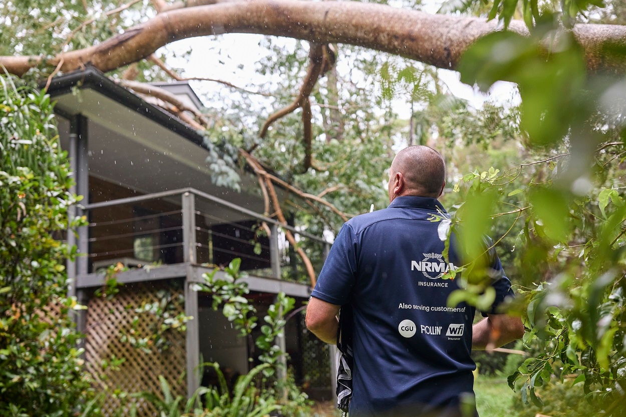 Rear view of a man in a dark blue NRMA Insurance branded shirt inspecting a house damaged by a fallen tree which is resting on the roof.