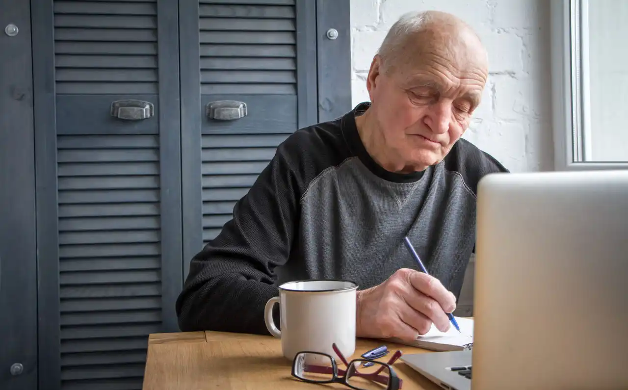 Older person sitting in front of laptop, writing on a notepad with glasses and a coffee cup on the table.