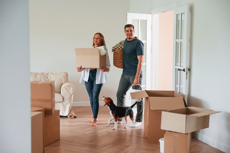 Photo of two people and a dog carrying boxes into a new house.