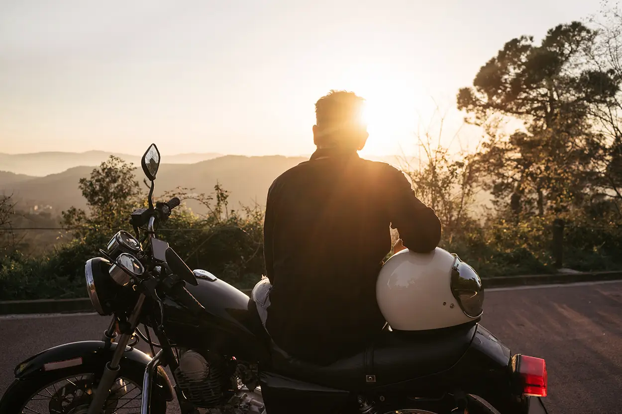 Back of a person sitting on the side of a motorcycle looking towards a sunset over a mountain range.