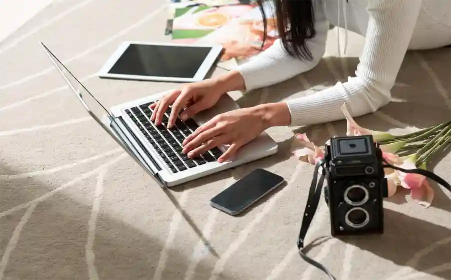 Person on Laptop with a tablet, phone and camera by their side lying down on a rug